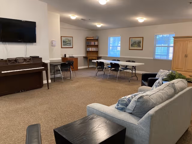 Community lounge with a sofa and armchair in the foreground, a piano and wall-mounted TV to the left, and folding tables and chairs by a bookshelf under windows.