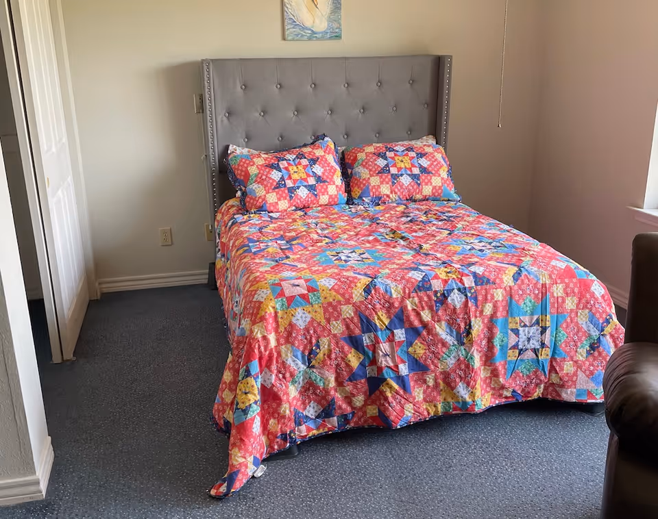 A bedroom with a bed featuring a colorful quilt and matching pillows with a star pattern. The bed has a gray tufted headboard. There is a small painting above the headboard, a window to the right, and a closet door to the left.