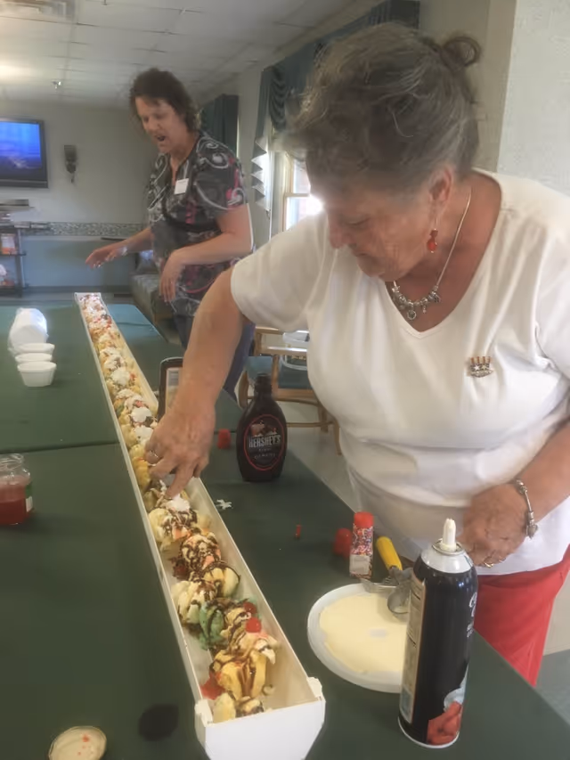 Two elderly women preparing and decorating a very long ice cream sundae on a green table in a communal room. One woman is adding toppings while the other stands in the background. Various ice cream toppings like chocolate syrup, whipped cream, and sprinkles are visible on the table.
