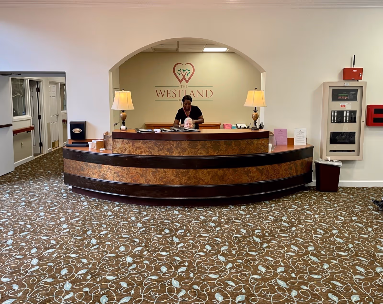 Reception area at The Westland with a curved wood front desk, a staff member behind it, two lamps, and patterned carpet.