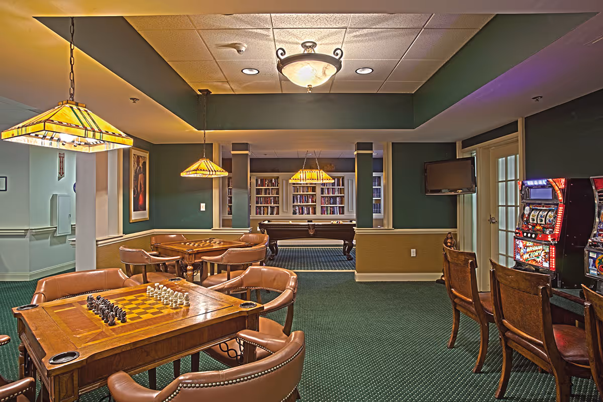 Interior recreation room with a chess/checker table and leather chairs in the foreground, hanging lamps, a pool table and bookshelves in the back, and slot machines to the right.