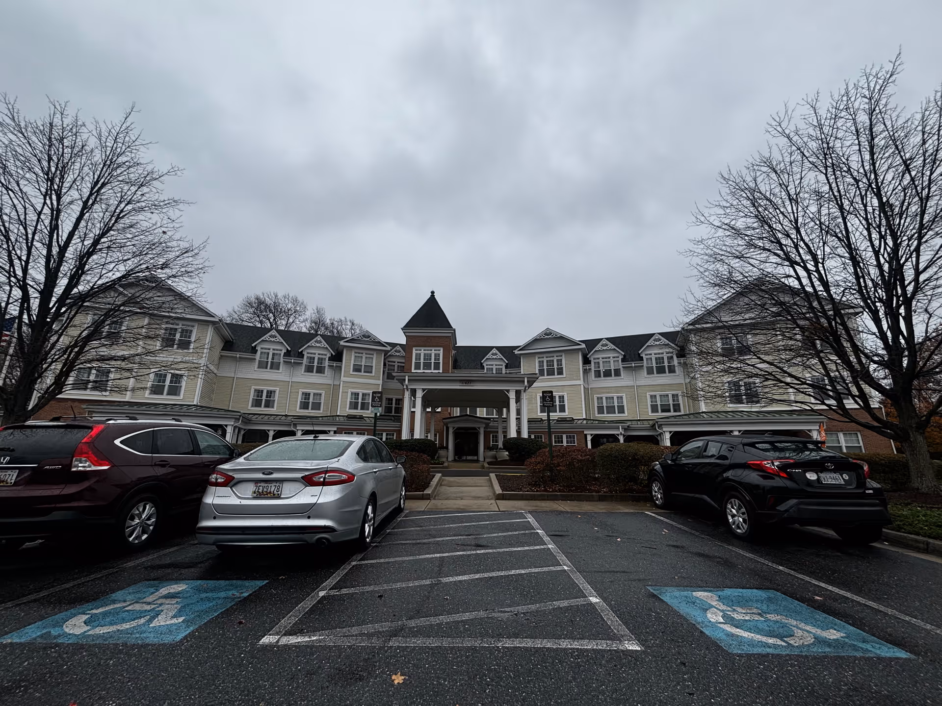 Front exterior of a multi-story senior living facility with a covered entrance, parked cars, and an overcast sky.