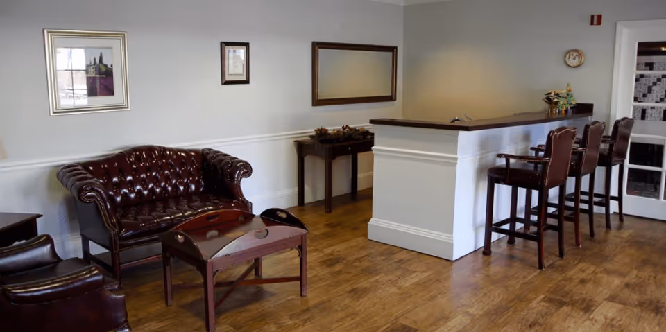 A cozy interior room with wooden flooring featuring a dark brown leather loveseat and armchair, a wooden coffee table, a small console table with decorations, a white half-wall counter with a dark wooden top, and three high-backed bar stools. The walls are light-colored with framed pictures and a clock, and a door with glass panels is visible in the background.