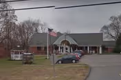 Exterior view of a single-story brick building with a gray roof, an American flag on a flagpole in front, several parked cars, and a small white gazebo on the lawn to the left.
