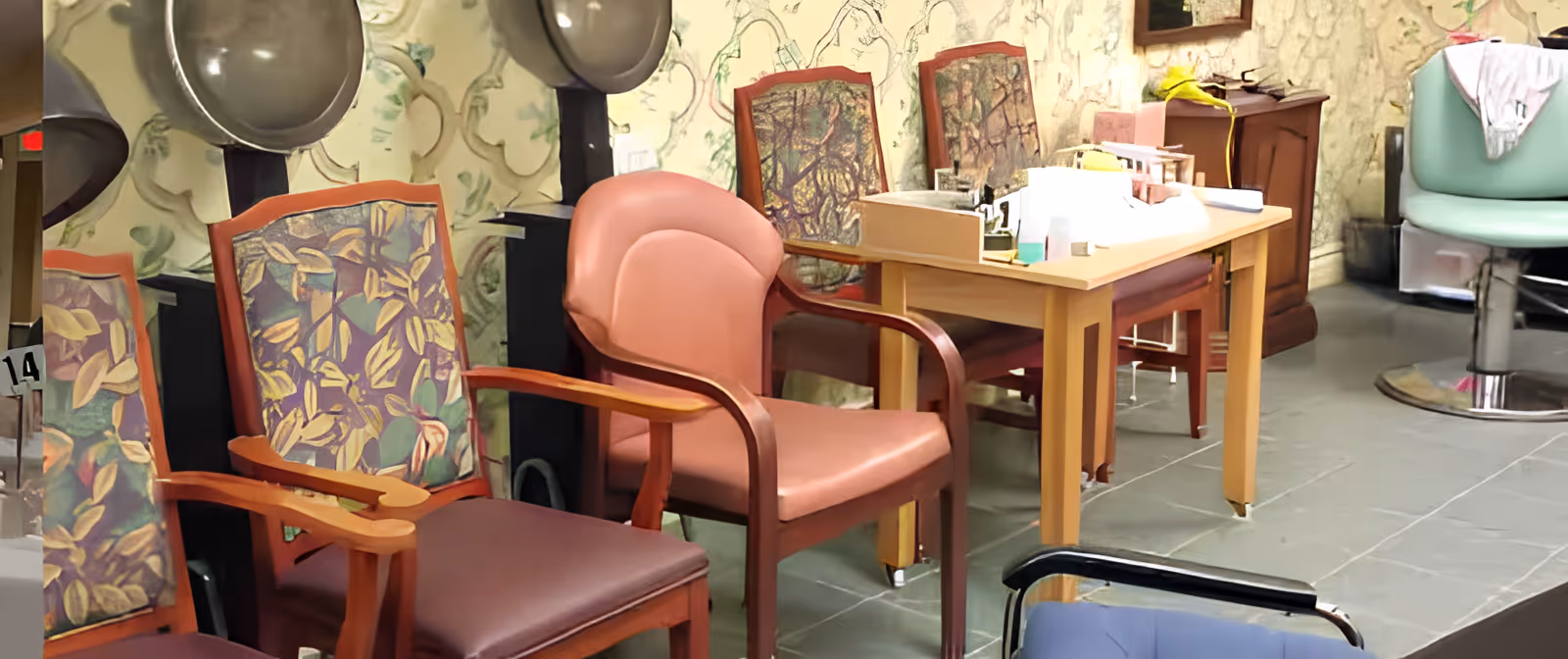 Interior view of a salon area with several chairs including floral patterned wooden chairs, a pink cushioned chair, and a green salon chair. There are two hair dryer hoods mounted on black stands against a wall with a decorative pattern. A wooden table with various items on it is also visible.