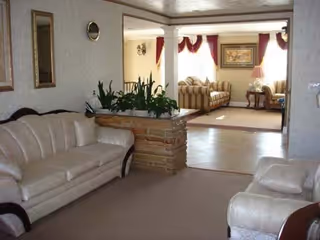 Interior view of a senior living facility showing a living room area with beige leather sofas, a brick planter with green plants, and a separate sitting area in the background with striped sofas, a side table with a lamp, and framed artwork on the wall.