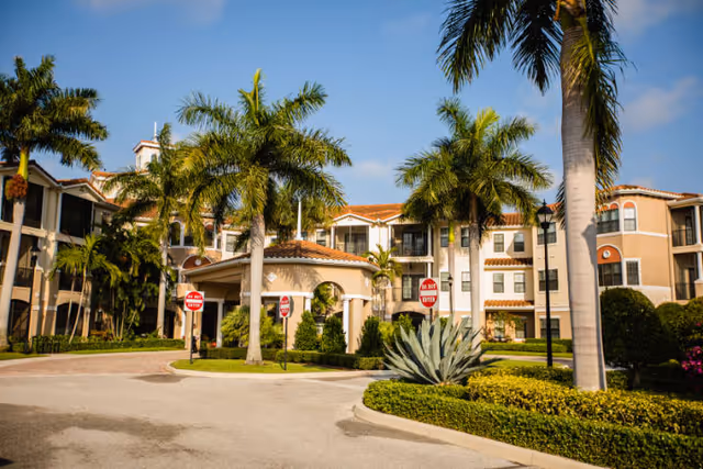 Exterior view of a senior living facility with a circular driveway, palm trees, and a covered entrance. The building has a Mediterranean architectural style with beige walls and red-tiled roofs under a blue sky.