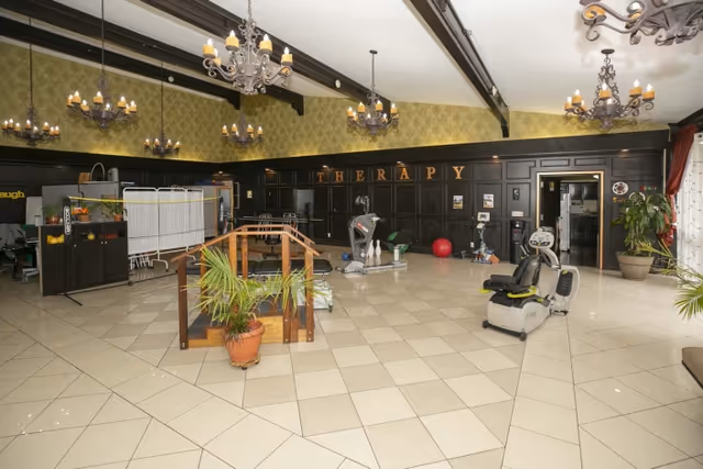 Spacious rehabilitation therapy room with exercise equipment, parallel bars, potted plants, and hanging chandeliers.