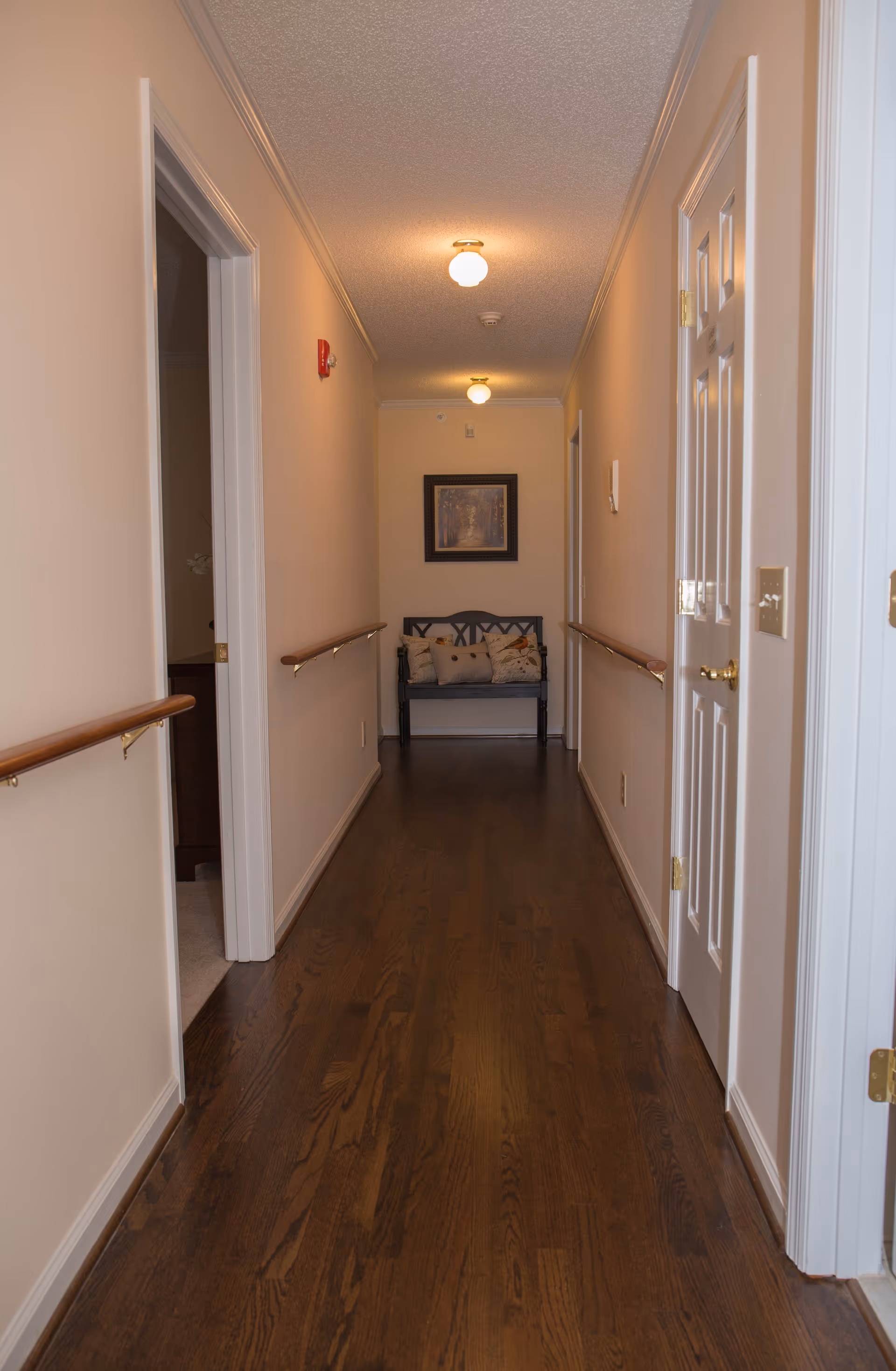 A long, narrow hallway with wooden flooring and beige walls. There are handrails on both sides of the hallway. At the end of the hallway, there is a dark wooden bench with several decorative pillows and a framed picture hanging above it. The hallway is lit by ceiling lights.