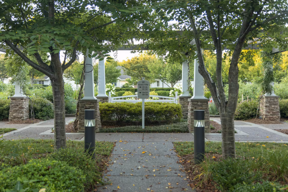 A tree-lined paved walkway leads to a pergola and landscaped courtyard with stone columns and a central sign.