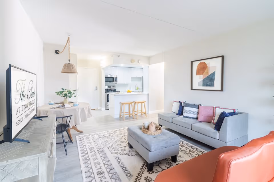 Bright and modern senior living facility interior featuring a living room with a gray sofa adorned with colorful pillows, a matching ottoman with decorative items, a patterned area rug, and an orange armchair. In the background, there is a small dining area with a round table and chairs, and a kitchen with white cabinetry, stainless steel appliances, and a breakfast bar with two wooden stools. A TV screen on a console displays the text 'The Park at Trowbridge Senior Living Community.'