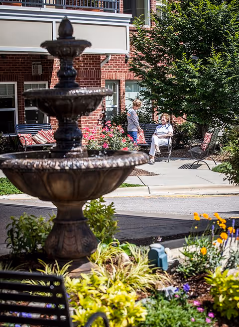Outdoor garden area at Preston Pointe featuring a multi-tiered water fountain in the foreground, surrounded by plants and flowers. In the background, two women are conversing near a brick building with benches and greenery around.