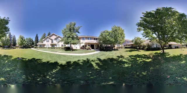 Wide panoramic view of the outdoor grounds of Marbella Chico facility featuring a large green lawn, several mature trees, and a two-story building with multiple windows under a clear blue sky.