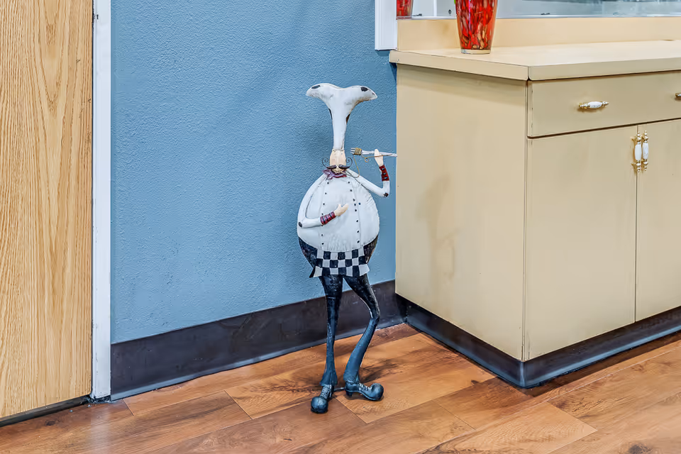 A whimsical metal sculpture of a chef with a tall hat, mustache, and checkered apron stands on a wooden floor next to a beige cabinet and a blue wall.