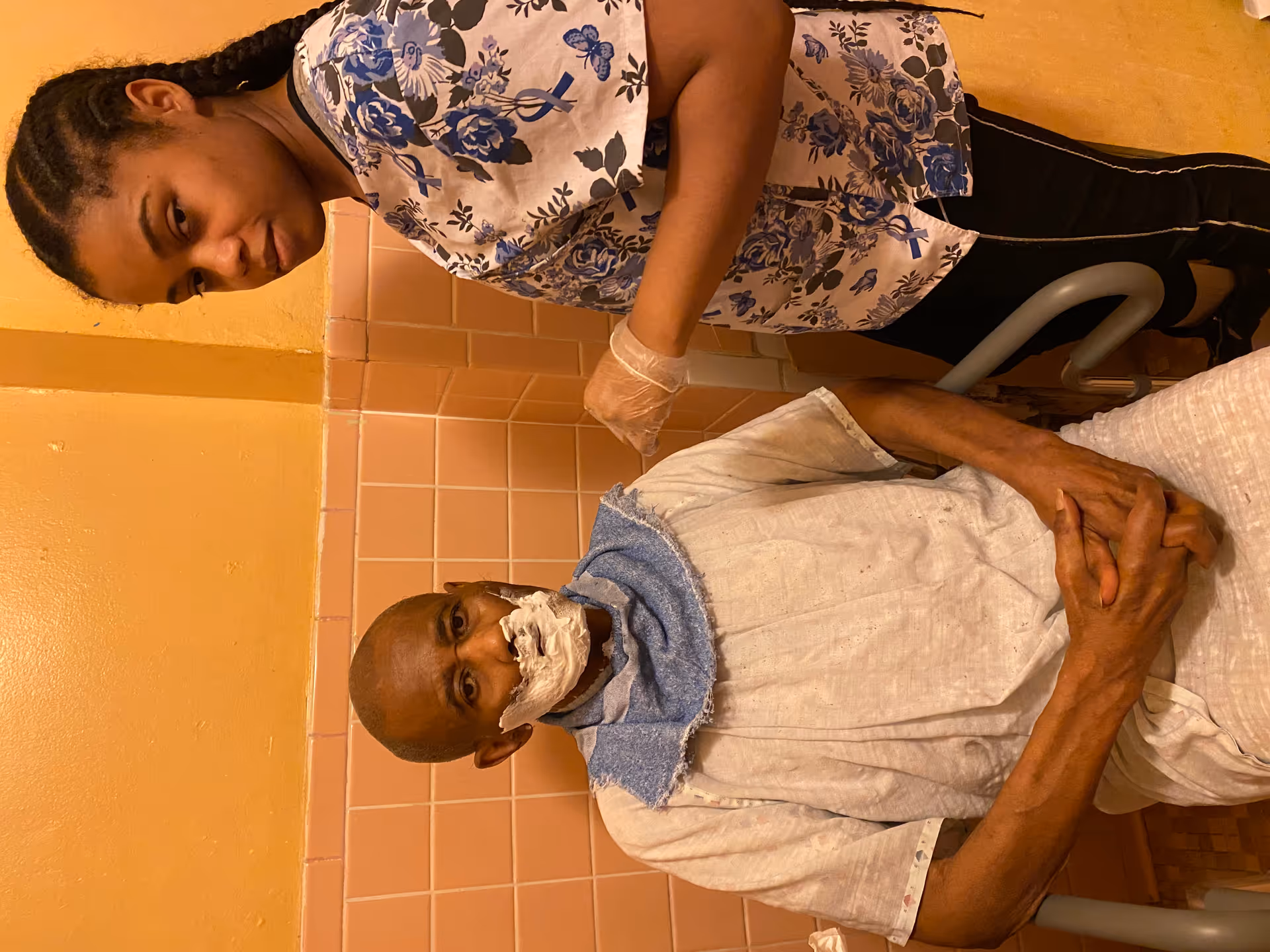 A caregiver stands beside an elderly person seated with shaving cream on their face in a pink-tiled room.