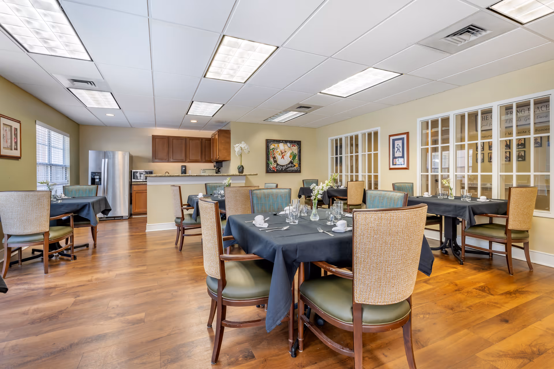 A dining room with several tables covered in black tablecloths, each set with glasses, napkins, and cutlery. The room has wooden flooring, beige walls, and a ceiling with fluorescent lights. There is a kitchen area in the background with wooden cabinets and a stainless steel refrigerator. The walls feature framed artwork and windows with white frames.