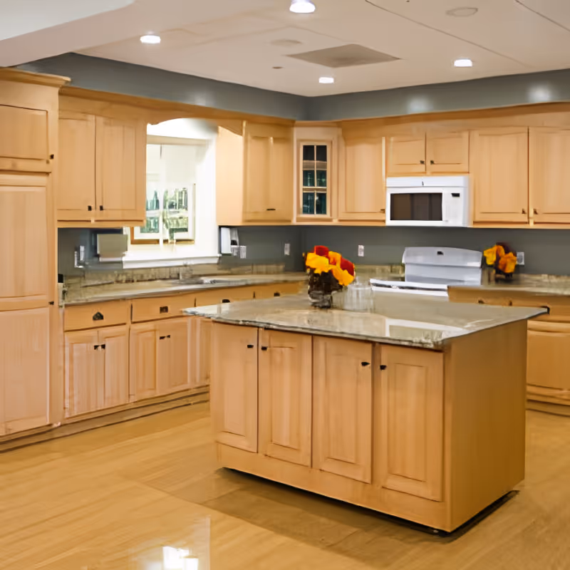 A spacious kitchen with light wood cabinetry and granite countertops. There is a central island with cabinets and a vase of yellow and red flowers on top. The kitchen includes a white microwave and stove, recessed lighting in the ceiling, and a window above the sink.