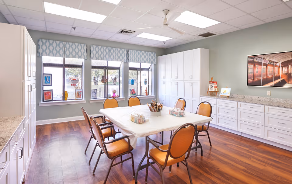 A bright and spacious activity room with a large table covered with a white cloth surrounded by six orange cushioned chairs. The room has wooden flooring, white cabinets along two walls, and three large windows with patterned valances letting in natural light. Various art supplies and small artworks are displayed on the table and windowsills.