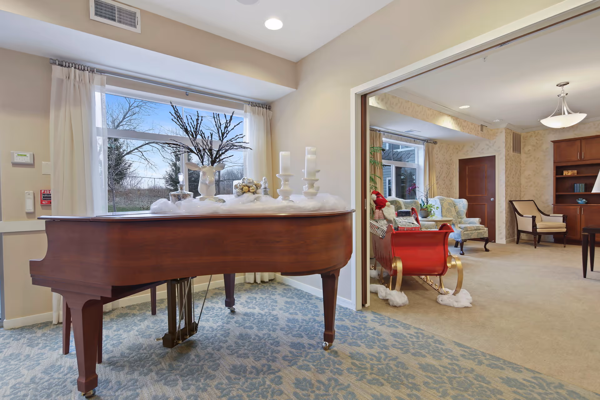 Interior view of a senior living facility room featuring a grand piano decorated with candles and holiday ornaments near a large window with white curtains. Adjacent room visible through an open doorway contains a red sleigh, armchairs, a wooden bookshelf, and a door, all set on carpeted floors.