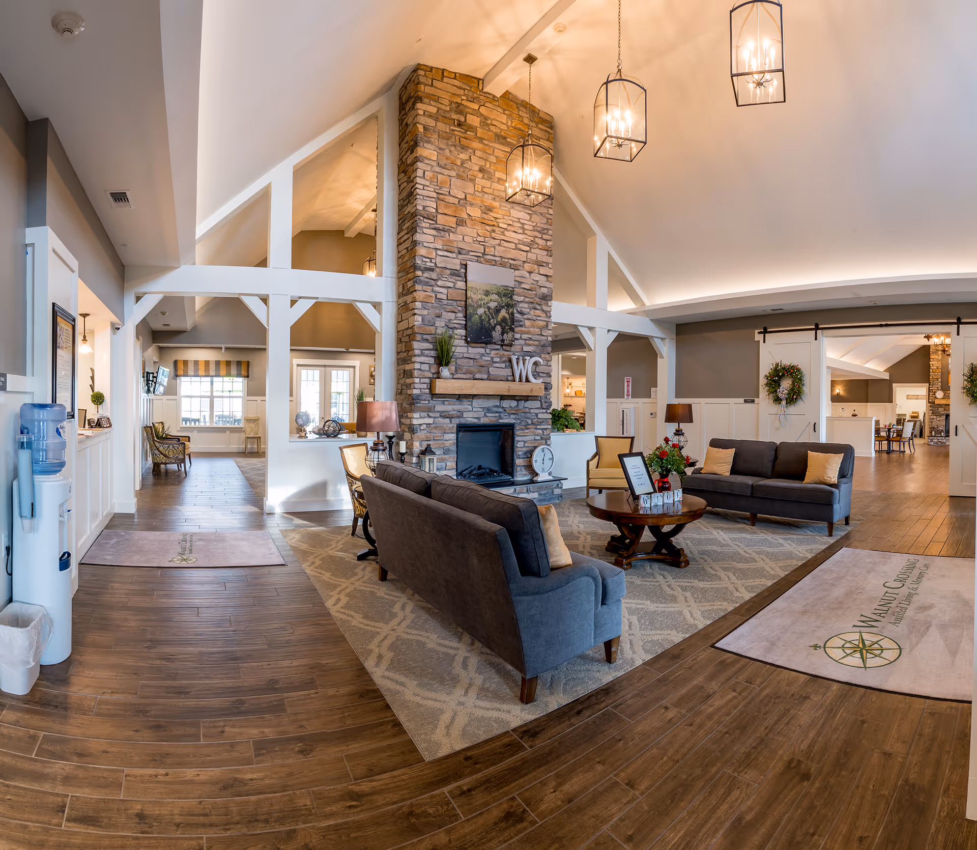 Spacious and well-lit living room area in Walnut Crossing facility featuring a stone fireplace with 'WC' letters mounted above it, two gray sofas with beige cushions, a round wooden coffee table with a flower arrangement, wooden flooring, and hanging lantern-style ceiling lights. The room has high vaulted ceilings with white beams and an open layout leading to other rooms.