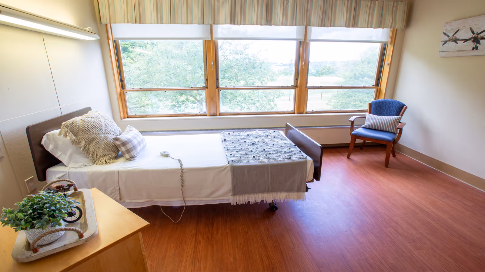 A bright and clean bedroom in a senior living facility with a single bed made up with white sheets, a textured pillow, and a gray throw blanket. There is a wooden nightstand with a potted plant and decorative tray. A blue armchair with a striped cushion is placed near large windows that let in natural light and offer a view of green trees outside. The room has wooden flooring and light-colored walls with a small airplane-themed wall decoration.