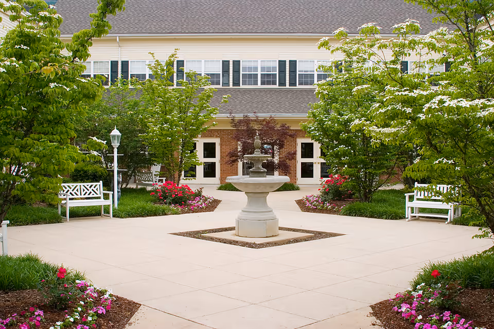 A landscaped outdoor courtyard area with a central stone fountain surrounded by paved walkways, flowering plants, green trees, white benches, and a building with multiple windows and doors in the background.