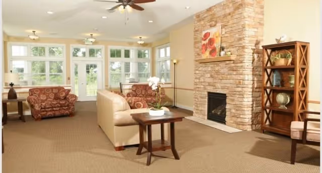 A spacious living room with beige walls and carpet, featuring a stone fireplace with a painting above it. The room is furnished with a beige sofa, two patterned armchairs, a wooden side table with a white orchid, a wooden bookshelf with decorative items, and large windows letting in natural light.