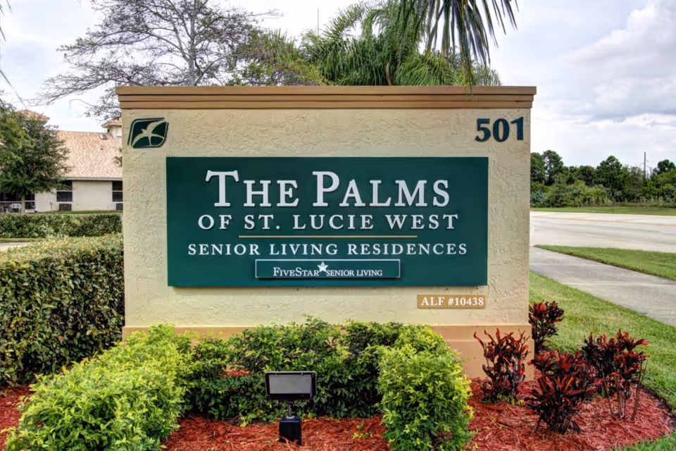 Outdoor sign for The Palms of St. Lucie West Senior Living Residences, surrounded by green bushes and red mulch, with a road and trees in the background.