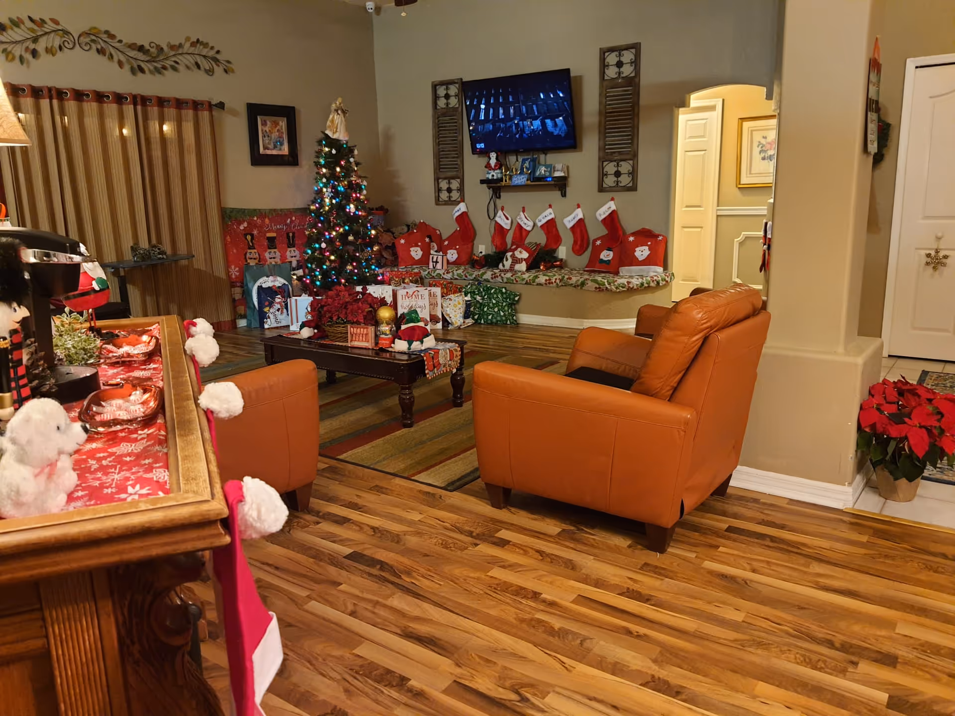 Cozy communal living room with leather armchairs, a decorated Christmas tree, stockings along a bench, and a wall-mounted TV.
