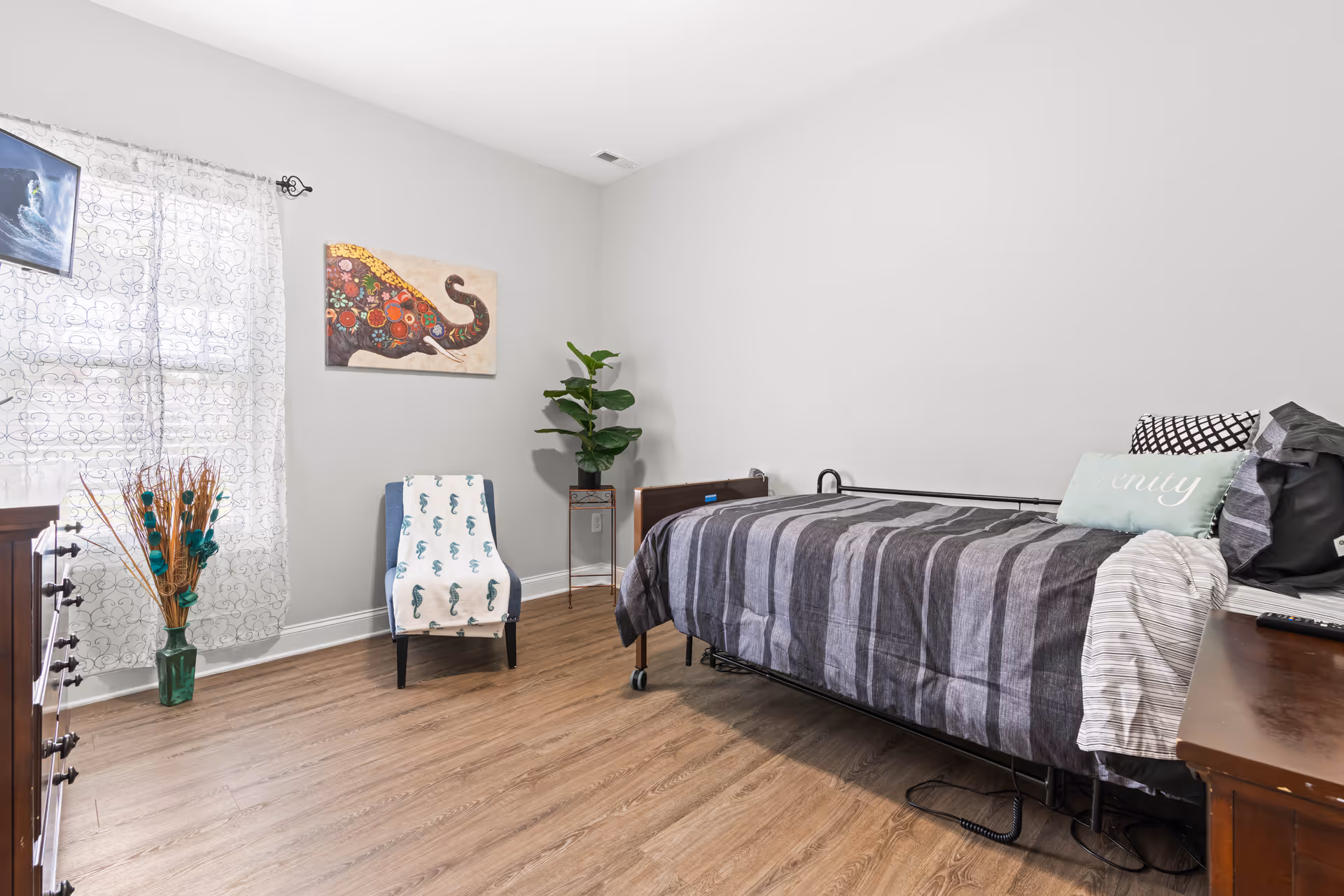Neat single bedroom with a bed, armchair, dresser, potted plant and elephant wall art.