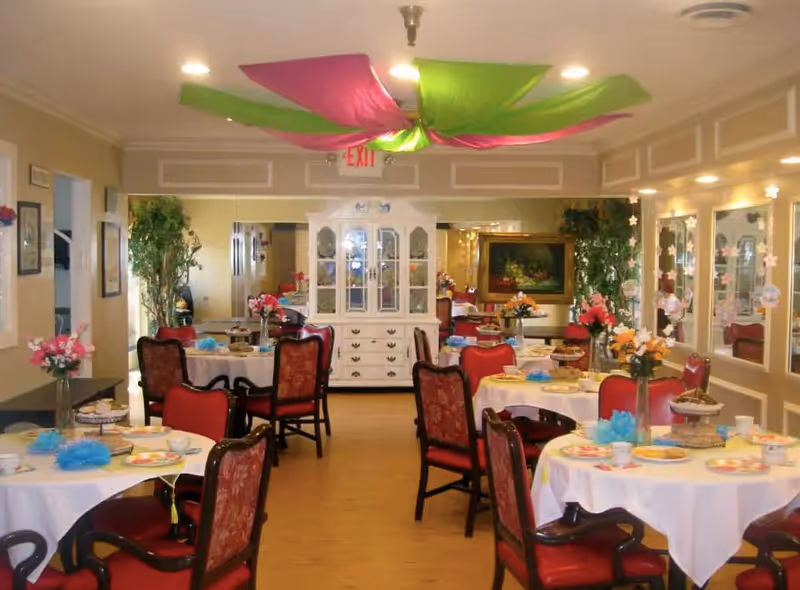 A dining room with round tables covered in white tablecloths, each set with plates, cups, and floral centerpieces. The room has red cushioned chairs with wooden frames, decorative plants, and a white cabinet against the far wall. The ceiling is decorated with pink and green fabric draped in a flower-like pattern.