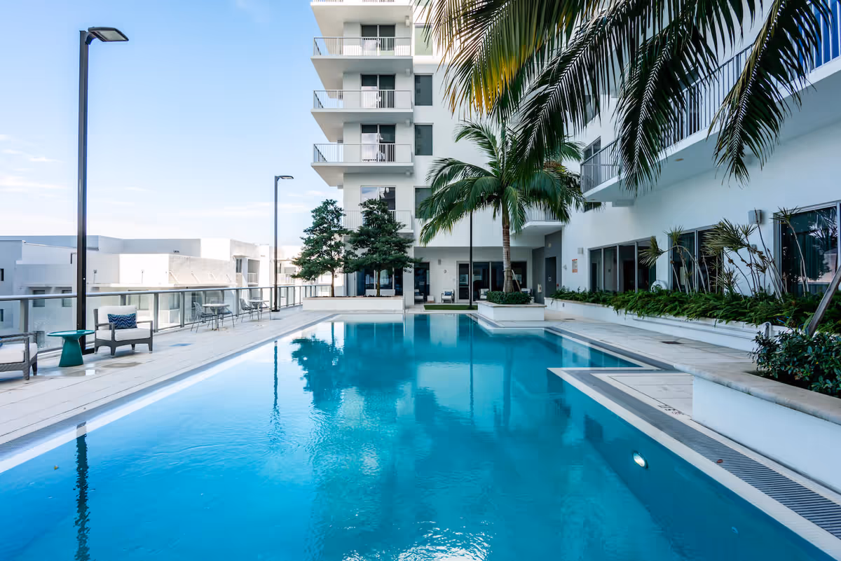 Outdoor swimming pool area at Mirabelle facility with clear blue water, surrounded by white buildings with balconies, palm trees, and seating areas with chairs and tables along the poolside.
