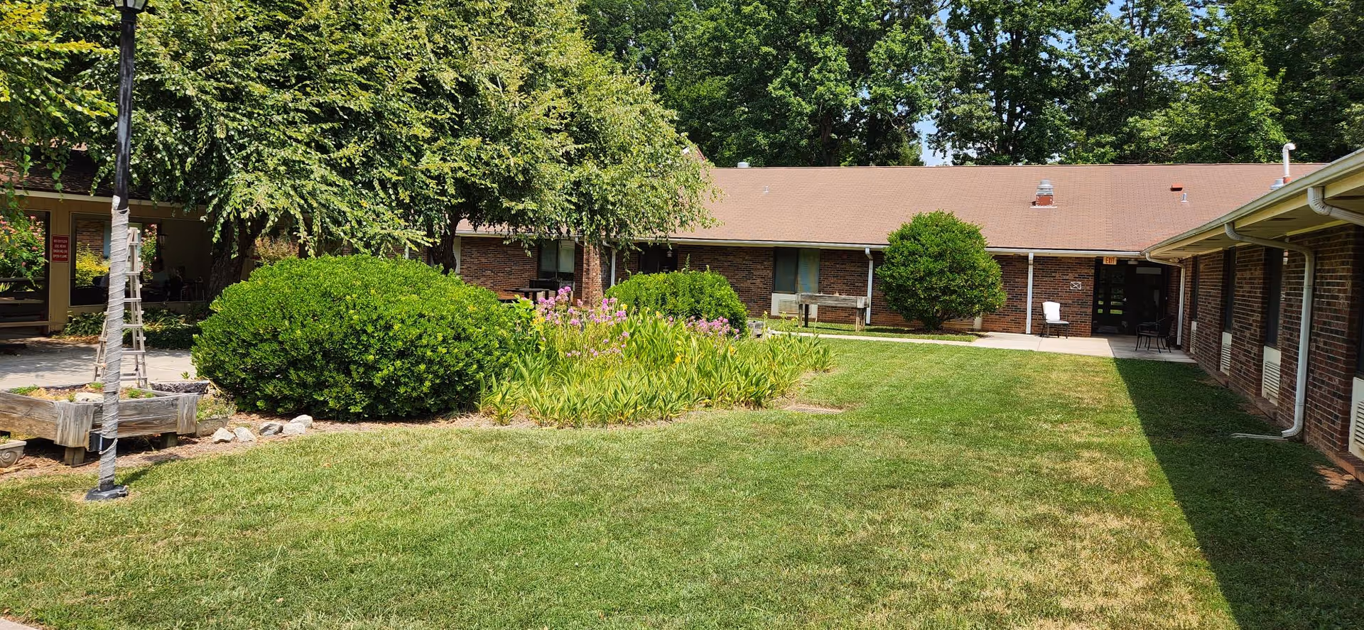 A grassy courtyard area surrounded by a single-story brick building with a brown roof. The courtyard features green bushes, flowering plants, and trees providing shade. There are benches and chairs placed near the building walls, and a paved walkway runs along the building edges.