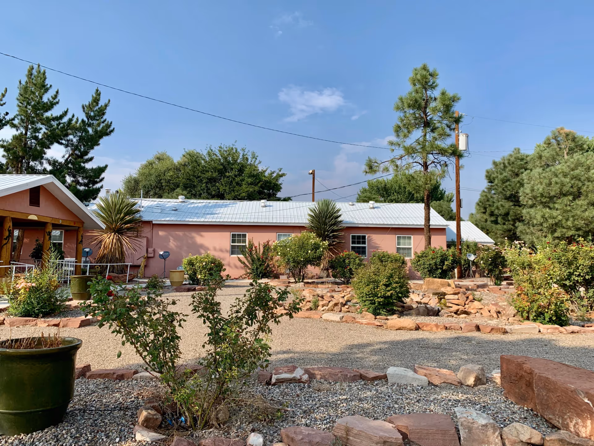 Outdoor view of Casa de Rosa Assisted Living showing a pink building with a white metal roof surrounded by a garden with various bushes, trees, and stone pathways under a clear blue sky.