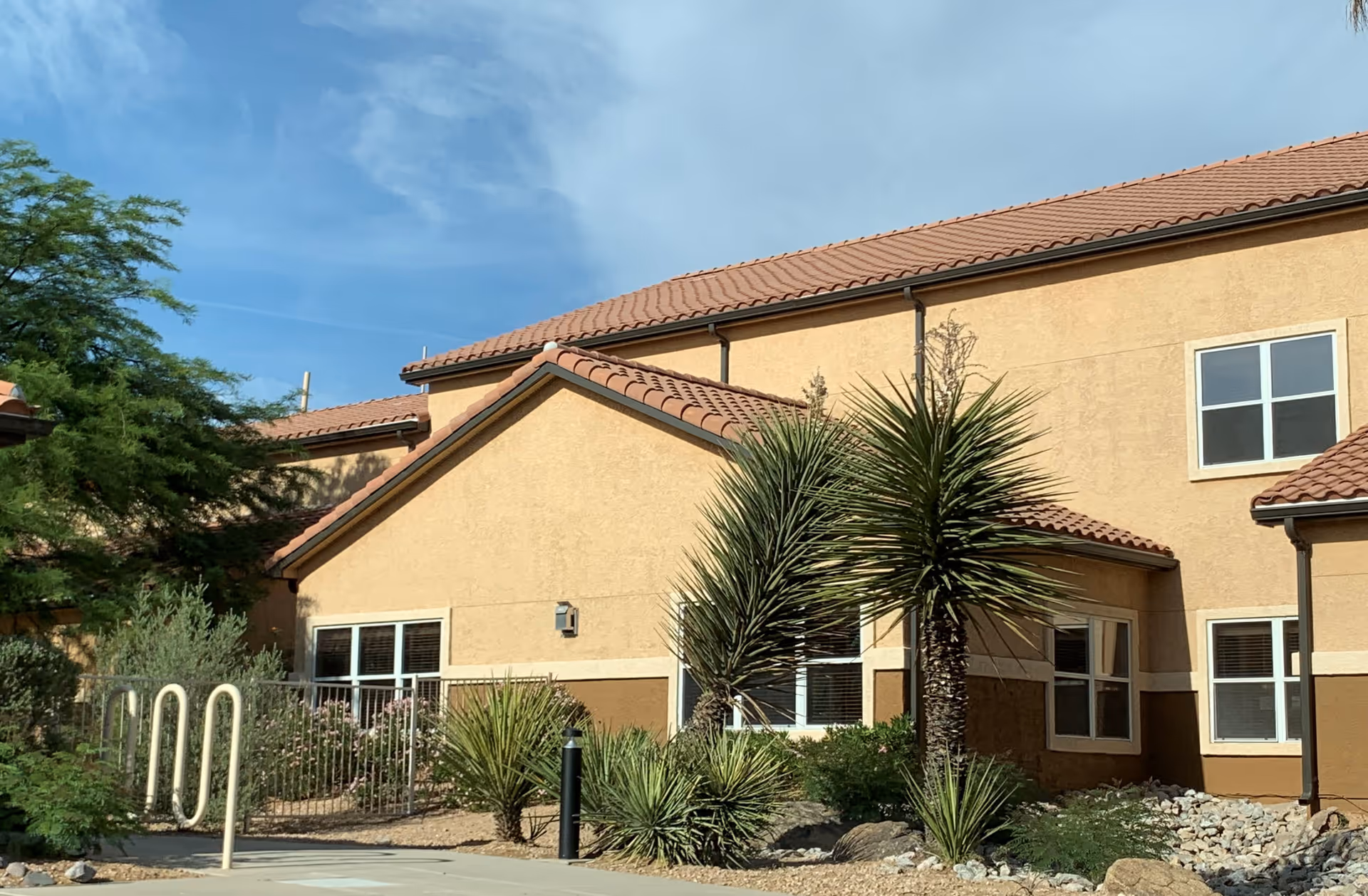 Exterior view of a beige stucco building with a red tile roof, surrounded by desert landscaping including palm-like plants and rocks, under a blue sky with some clouds.