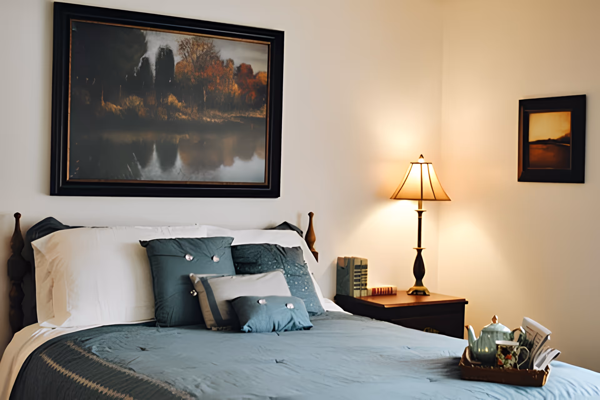 Well-lit bedroom with a neatly made bed dressed in teal bedding and decorative pillows, a nightstand with a lamp, and framed artwork on the walls.