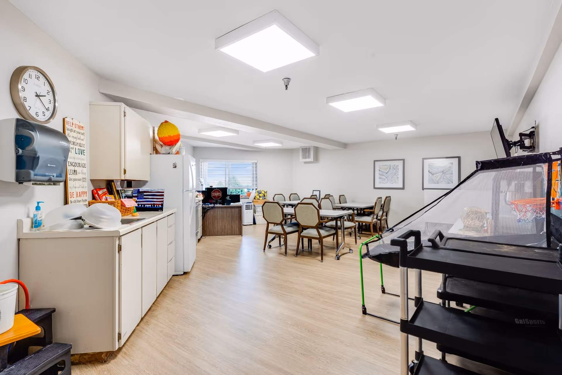 Bright communal activity and dining room with a kitchenette, several tables and chairs, and an indoor basketball arcade game.