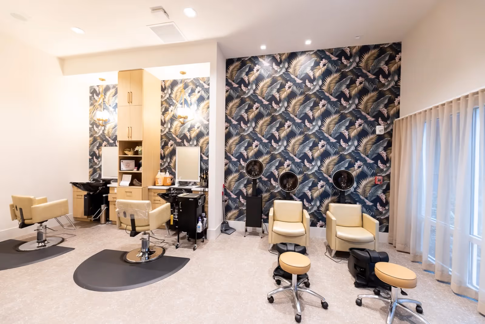Interior view of a salon area in Parasol Melbourne facility featuring two beige salon chairs with black hair washing sinks and mirrors on the left side, and two beige chairs with hair dryers and foot baths on the right side. The back wall has a dark floral and leaf patterned wallpaper, and there are large windows with sheer curtains on the right.
