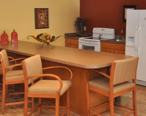 A kitchen area with a long countertop and three wooden chairs with cushioned seats. The kitchen features a white stove, a white refrigerator, wooden cabinets, and a decorative fruit bowl on the counter. The walls are painted in warm tones with a framed picture and a decorative plant in the corner.