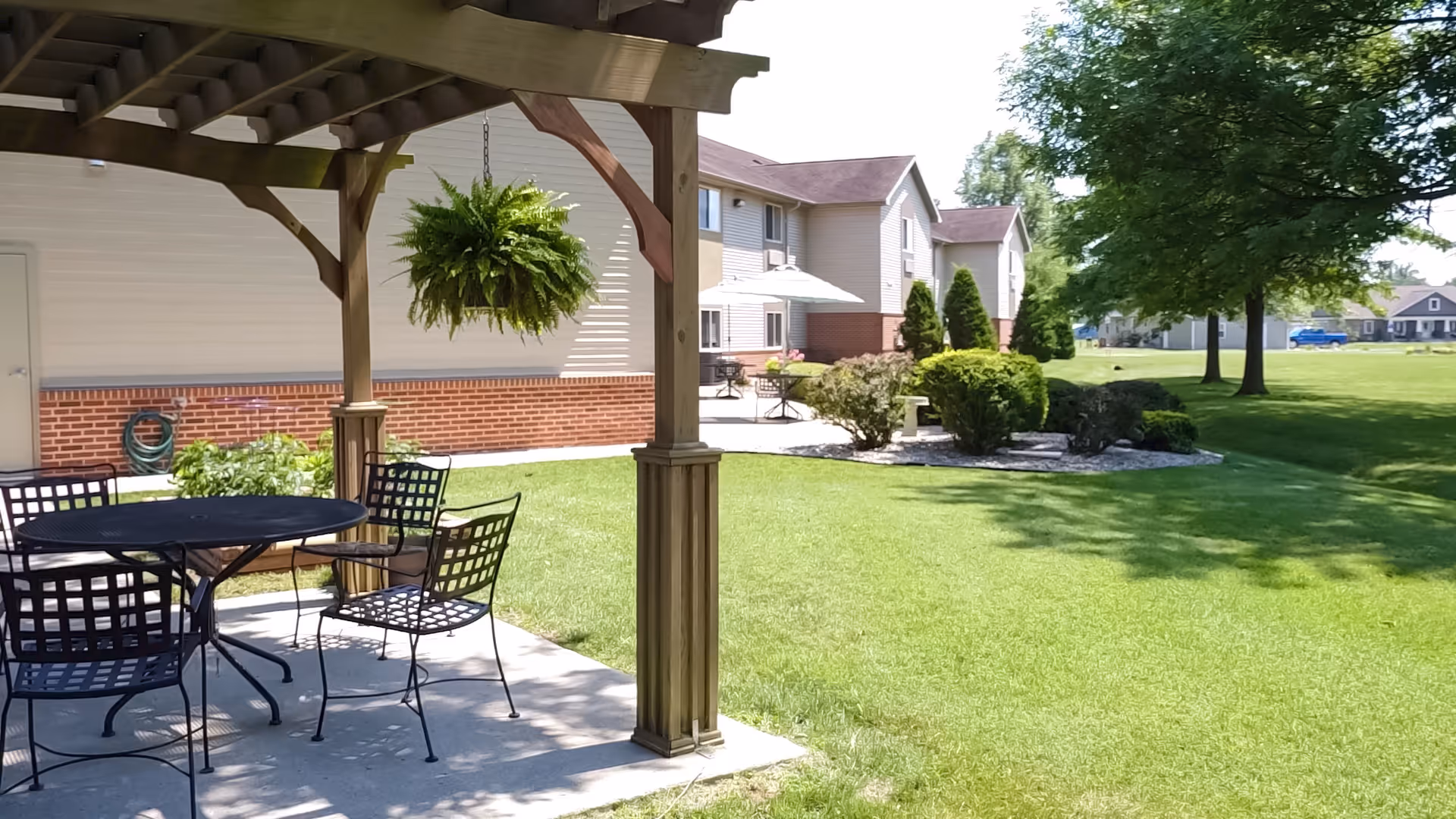 Outdoor patio area at Heritage Woods of Flora featuring a wooden pergola with a hanging fern plant, a round metal table with four matching chairs on a concrete slab, well-maintained green lawn, shrubs, trees, and residential buildings in the background under a sunny sky.