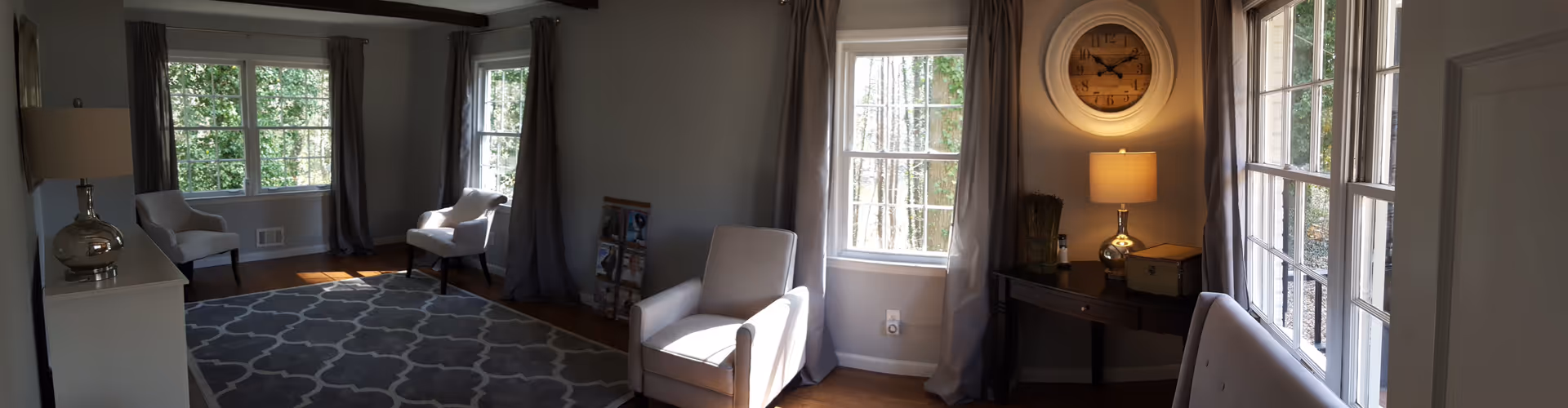 A cozy living room with multiple large windows letting in natural light, featuring several upholstered armchairs, a patterned area rug, a small black desk with a lamp and decorative items, and a large wall clock above the desk. The walls are painted light gray and the windows have gray curtains.