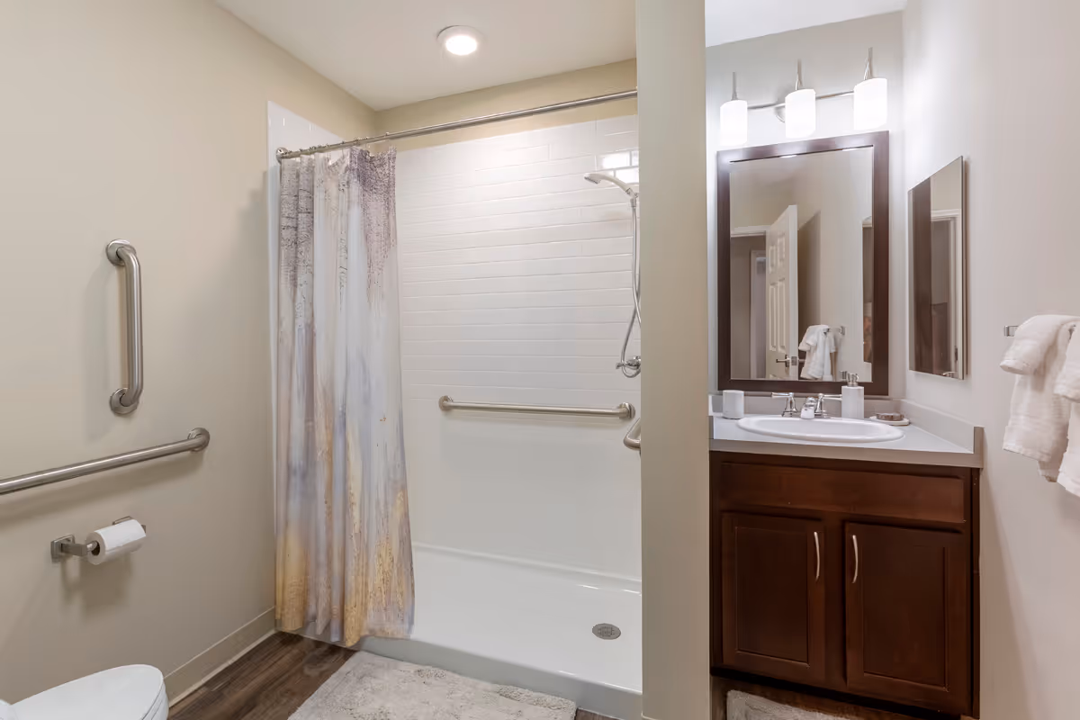 A clean and modern bathroom featuring a walk-in shower with a curtain, grab bars on the walls, a toilet with a toilet paper holder, and a vanity with a sink, mirror, and three light fixtures above. Towels are hanging on a rack on the right wall.