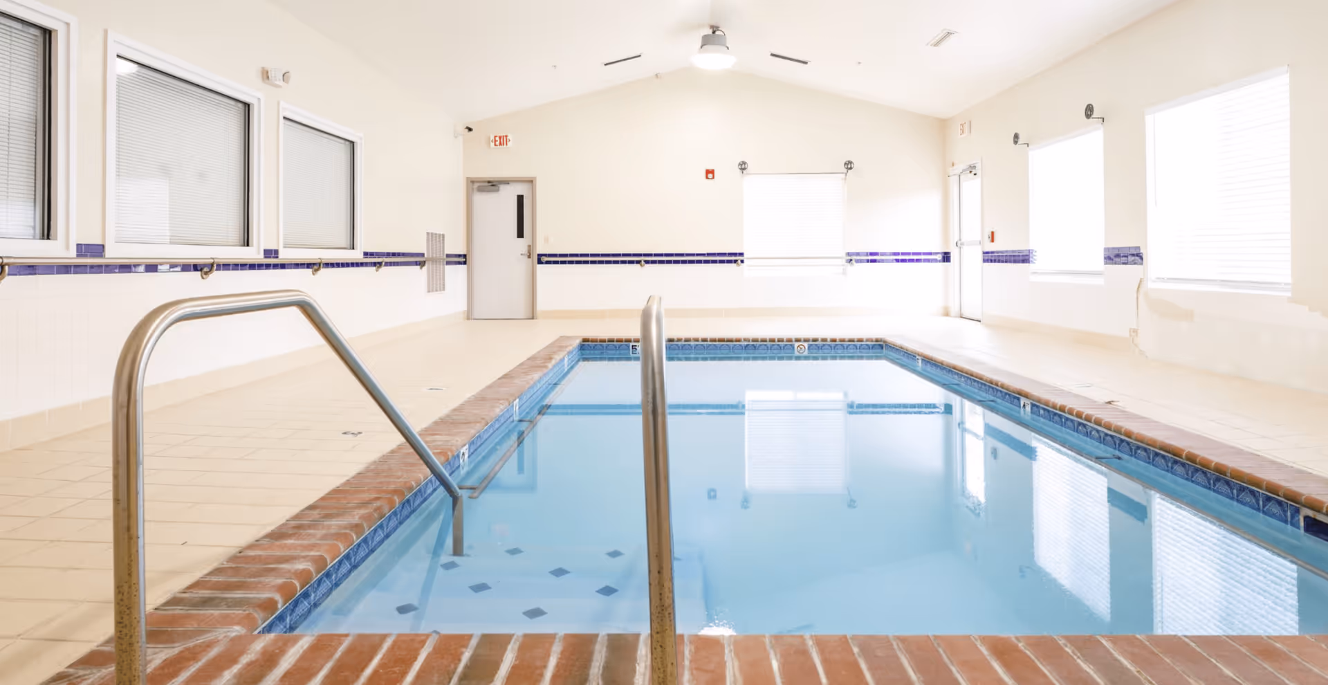 Indoor swimming pool with metal handrails and tiled deck in a bright, windowed room.