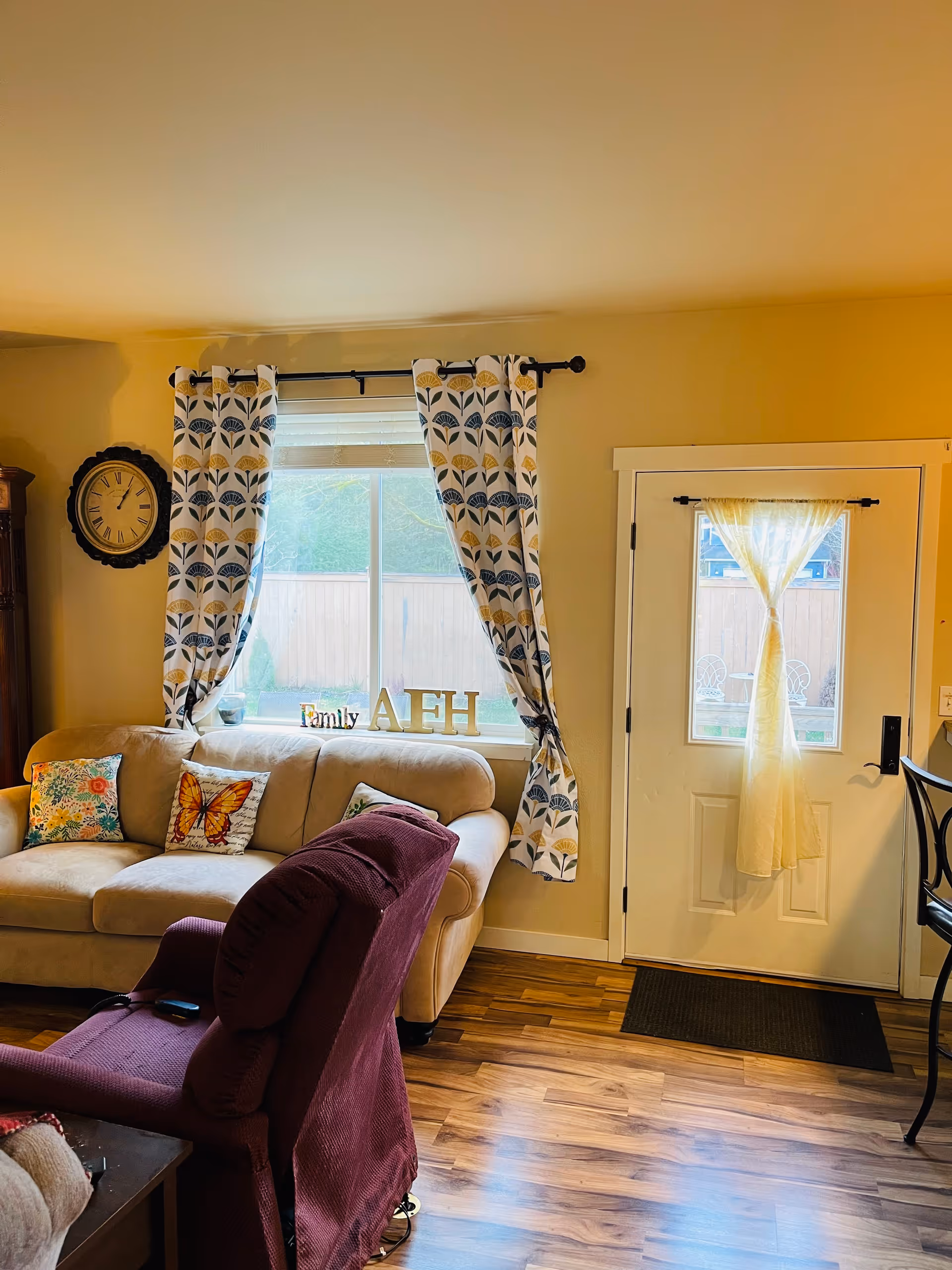 A cozy living room with a beige sofa adorned with colorful pillows, including one with a butterfly design. A maroon recliner chair is positioned in front of the sofa. Behind the sofa is a window with patterned curtains and decorative letters spelling 'Family AFH' on the windowsill. To the right is a white door with a sheer curtain tied in the middle, and a black mat on the wooden floor.
