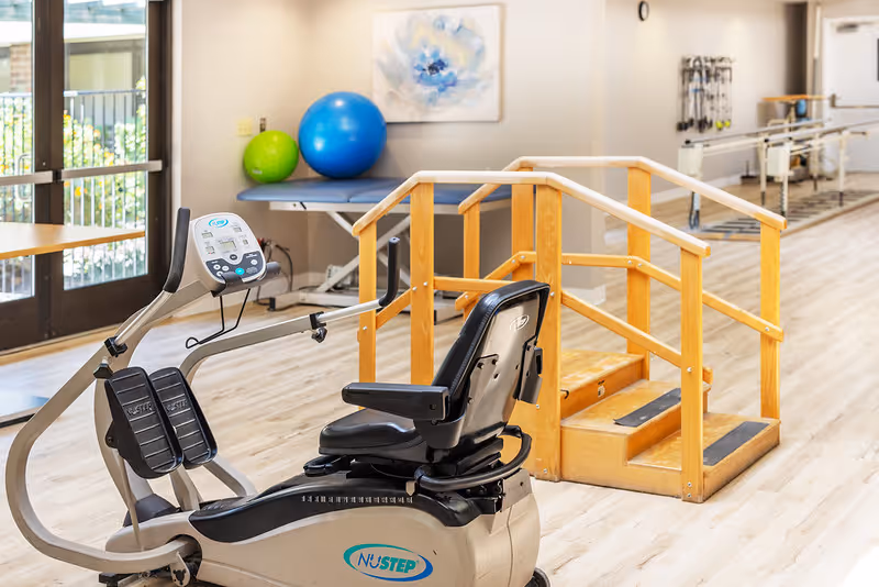 A physical therapy room with a NuStep exercise machine in the foreground, wooden handrails around a small set of stairs, exercise balls on a table, and various therapy equipment in the background.