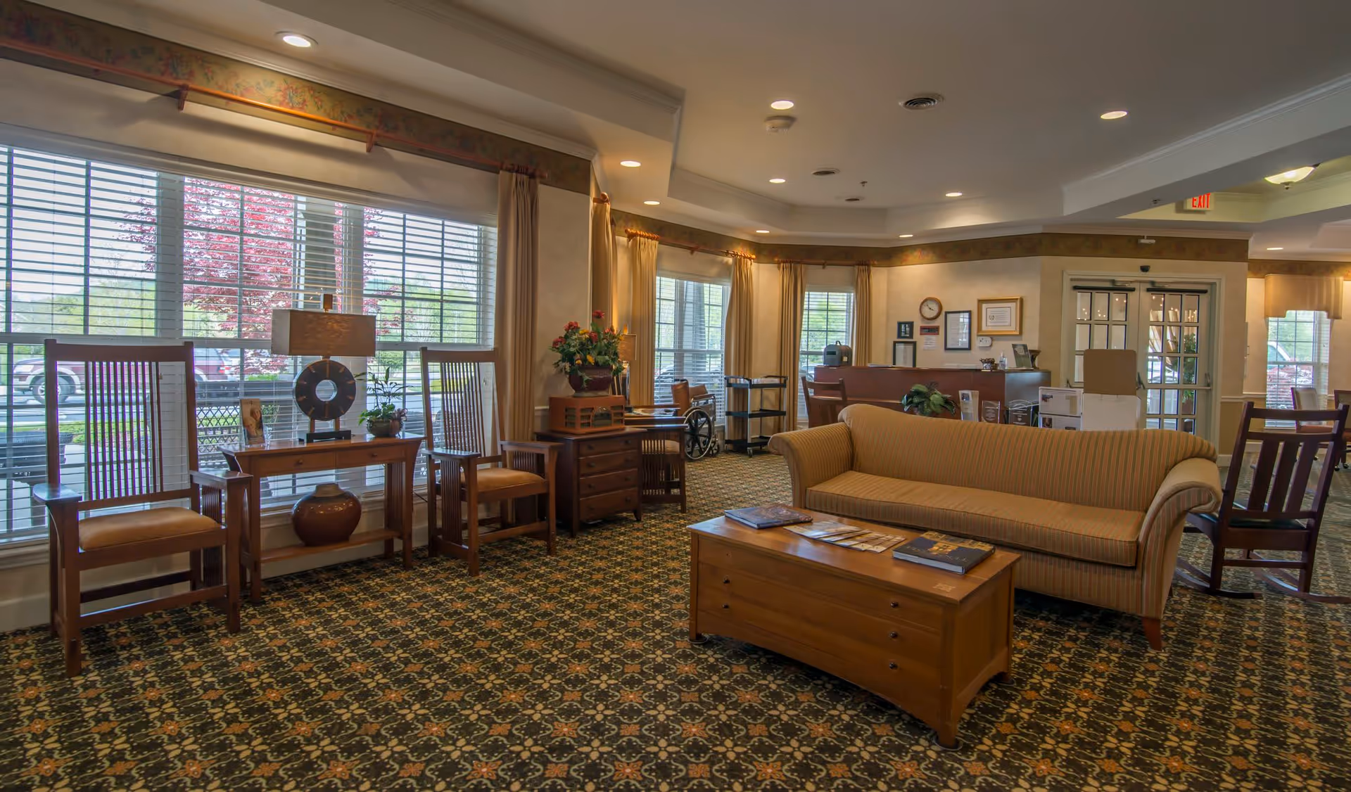Spacious, well-lit senior living lounge with sofas, wooden chairs, a coffee table, and large windows overlooking the outside.