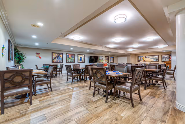 A spacious dining room with multiple wooden tables and chairs arranged neatly on a wood-patterned floor. The room is well-lit with ceiling lights and decorated with framed pictures on the walls. There is a large mirror and a television mounted on the far wall.