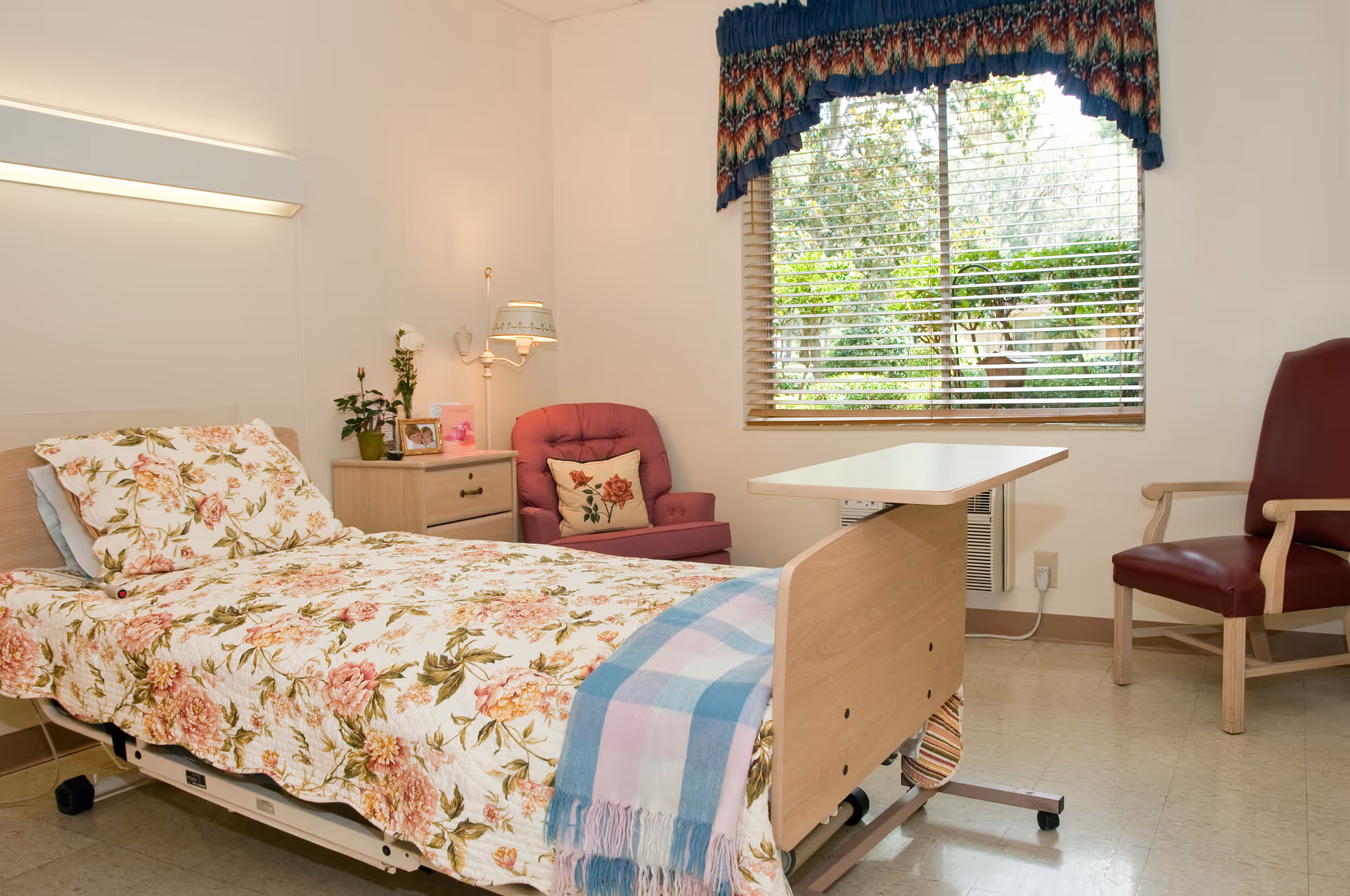 A bright senior care bedroom with a floral-covered hospital bed, overbed table, armchairs and a window with blinds.