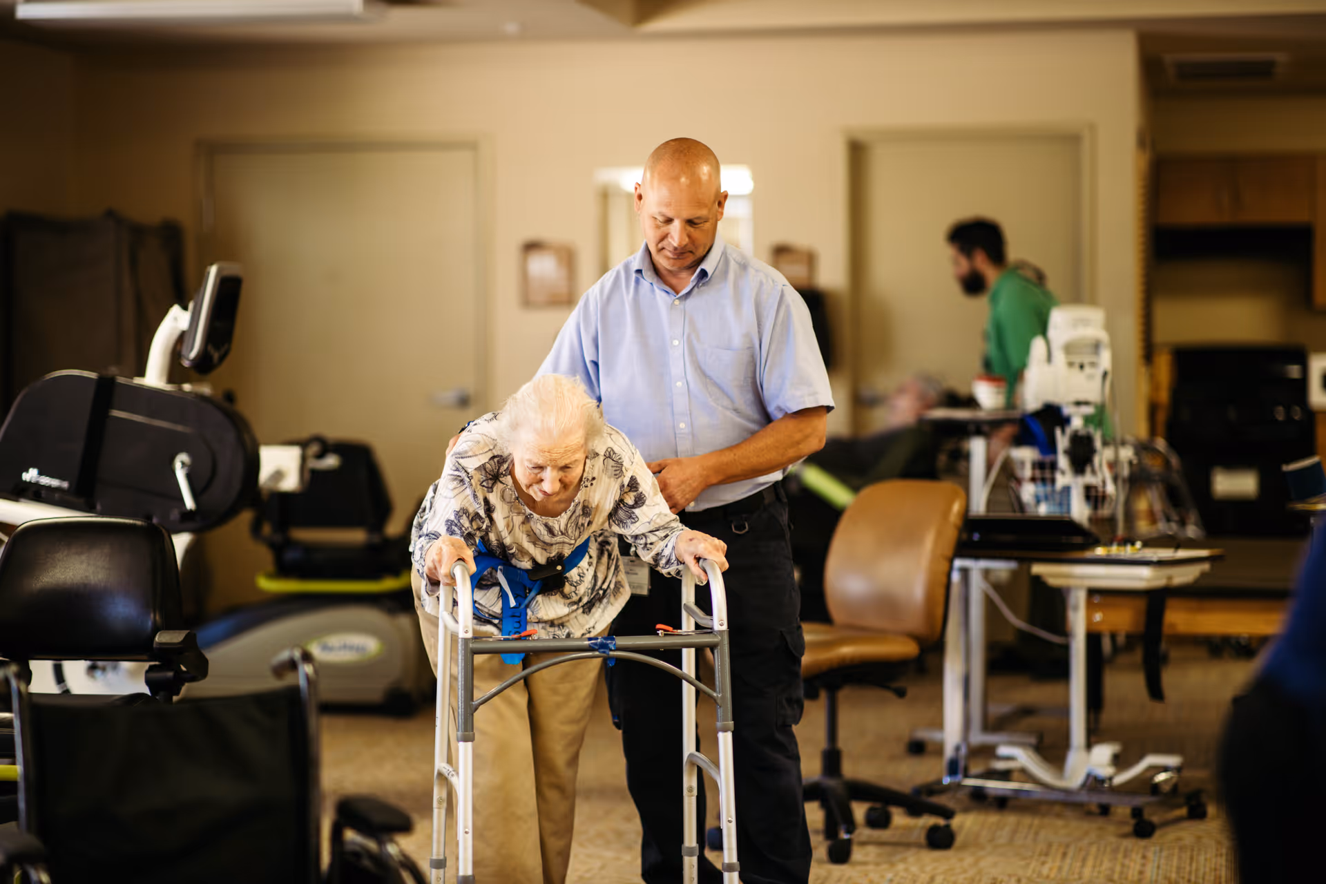 An elderly woman using a walker is assisted by a man in a light blue shirt in a room with medical and exercise equipment. Another person is visible in the background near medical devices and chairs.