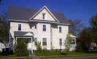 Two-story light-colored building with a gabled roof, front entrance, and surrounding trees.